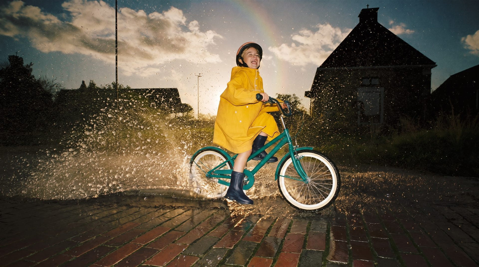 Kind fietst lachend door een grote plas, spatwater in gouden tegenlicht met een regenboog