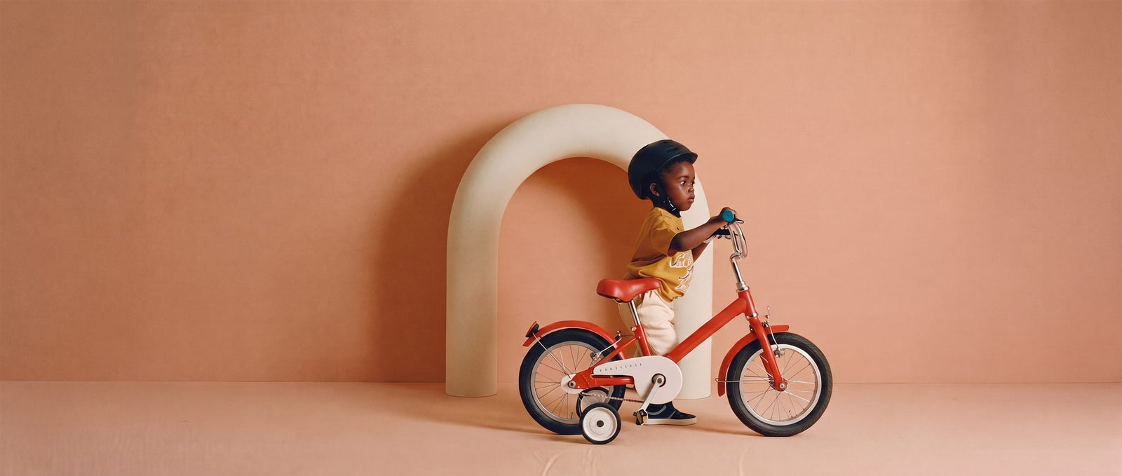 Peuter rijdt op een rood 12 inch fietsje met zijwieltjes voor een creme boog in een perzikkleurige studioset