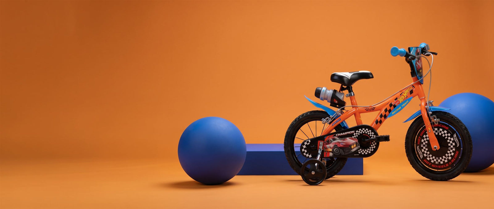 Oranje Dino Bikes kinderfiets met zijwieltjes in een oranje studioset met blauwe ballen