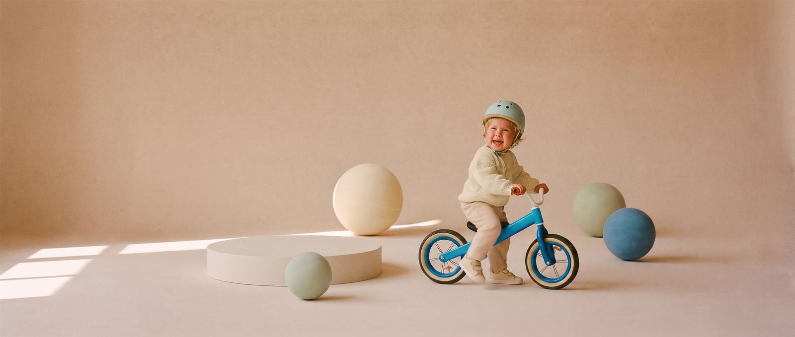 Lachende peuter zit op een blauwe loopfiets in een zachte perzikkleurige studioset met grote ballen