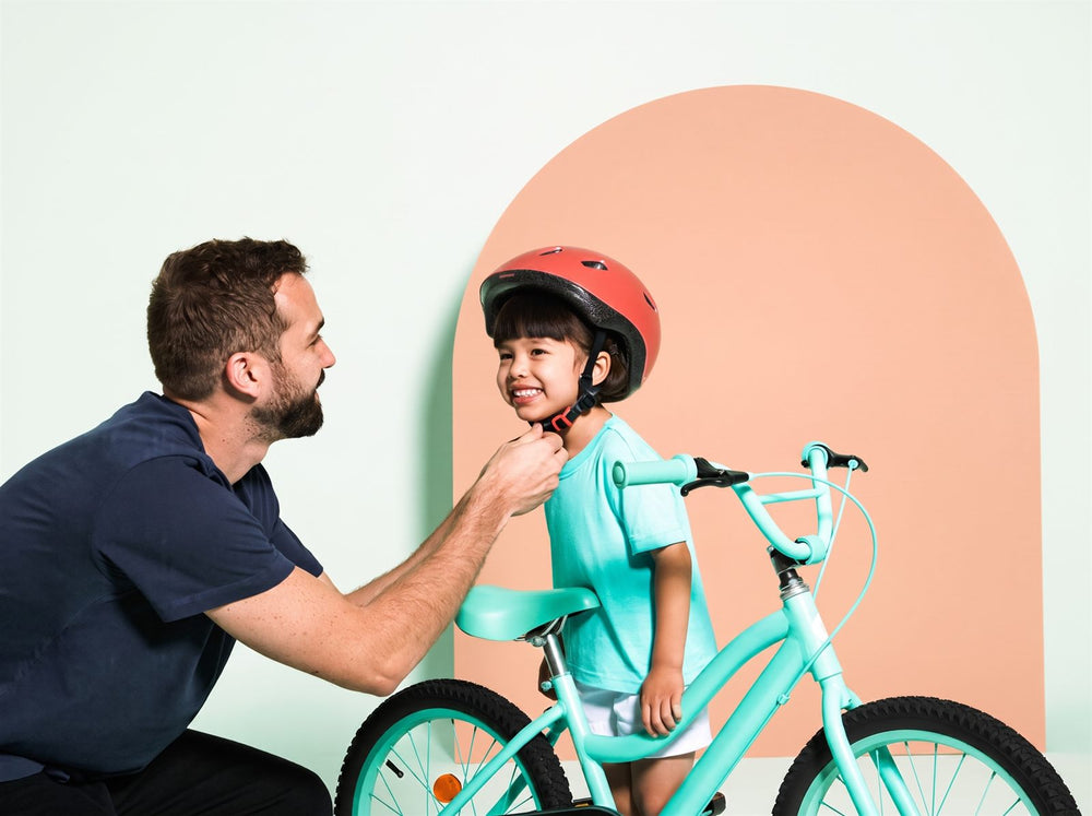 Een vader klikt de fietshelm vast van zijn dochter, naast haar staat haar kinderfiets