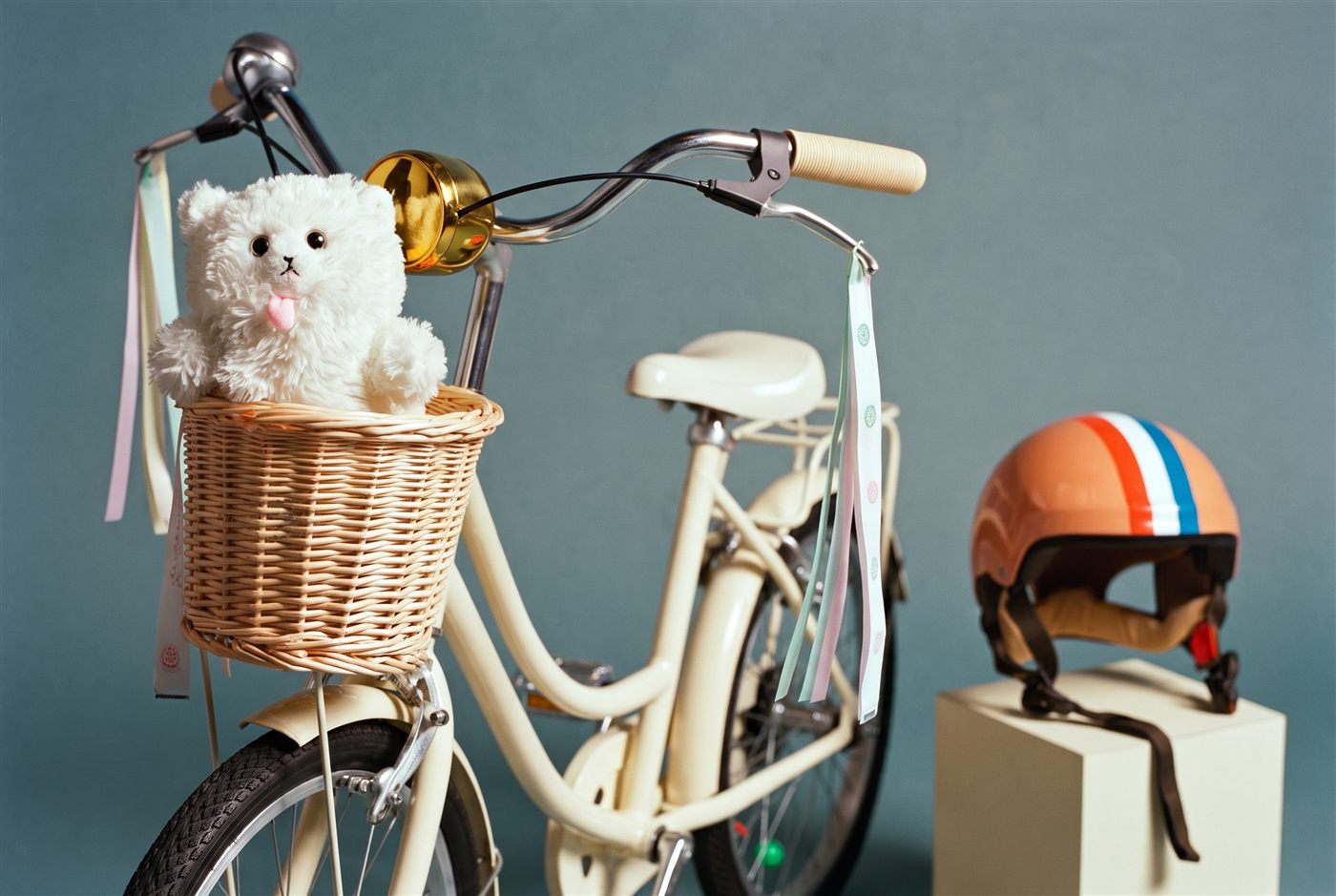 Close-up van een kinderfietsstuur met rieten mandje, knuffel, gouden bel en pastelkleurige linten in een teal studioset