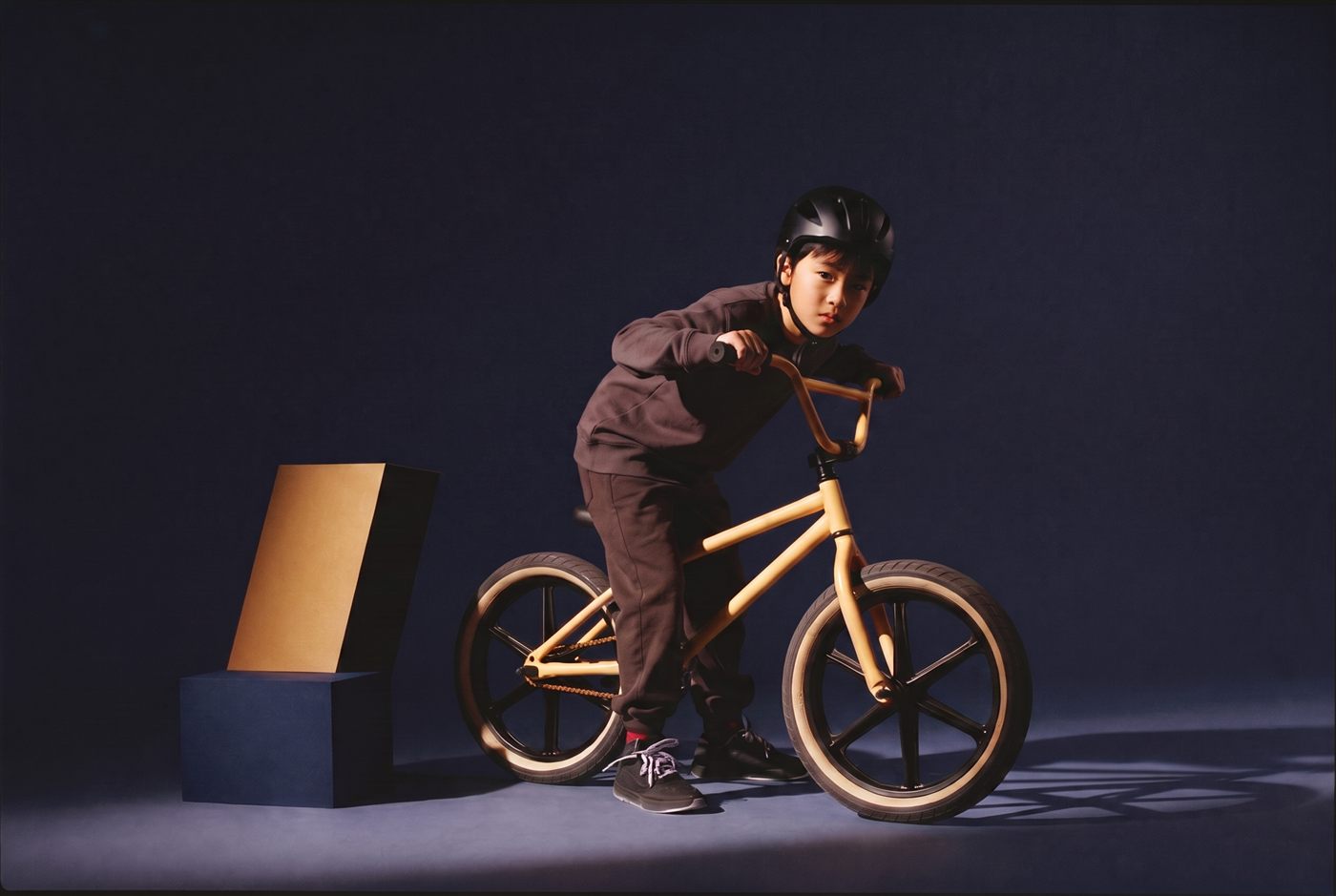 Stoere jongen leunt over het stuur van zijn zandgele BMX in een donkere navy studioset
