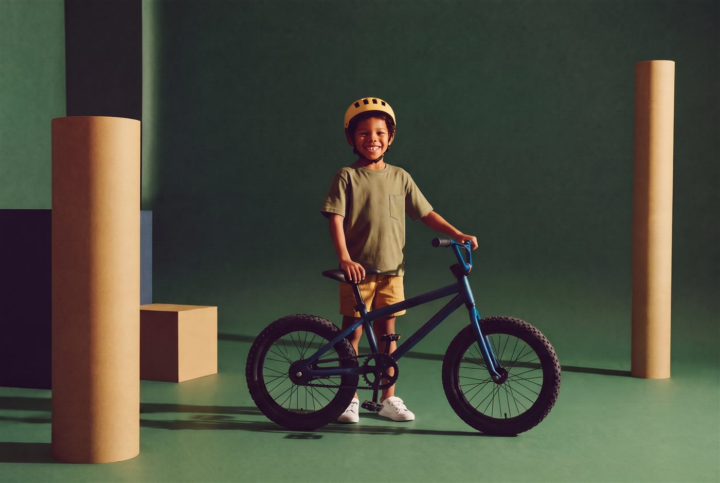 Jongen met gele helm staat trots naast zijn donkerblauwe fiets in een diepgroene studioset