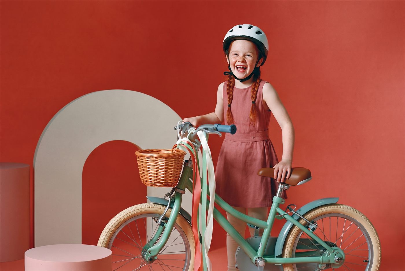 Meisje met rode vlechtjes staat lachend naast haar mintgroene fiets met rieten mandje in een koraalrode studioset