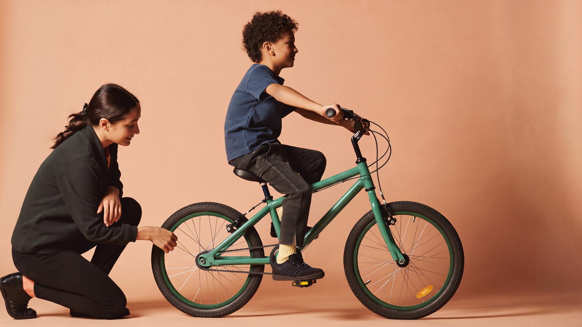 Moeder controleert lachend de pasvorm terwijl haar zoon op het zadel van een zeegroene kinderfiets zit in een perzikkleurige studio