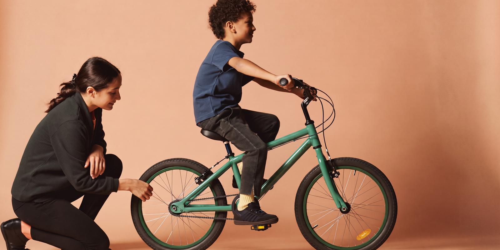 Moeder controleert lachend de pasvorm terwijl haar zoon op het zadel van een zeegroene kinderfiets zit in een perzikkleurige studio