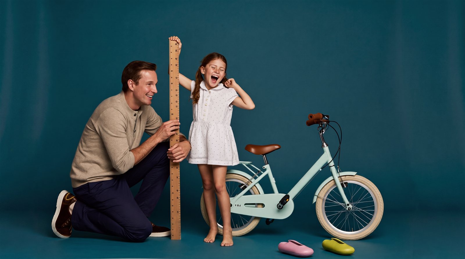 Vader meet lachend de lengte van zijn dochter bij een houten meetlat, naast een mintgroene kinderfiets in een zeegroene studio