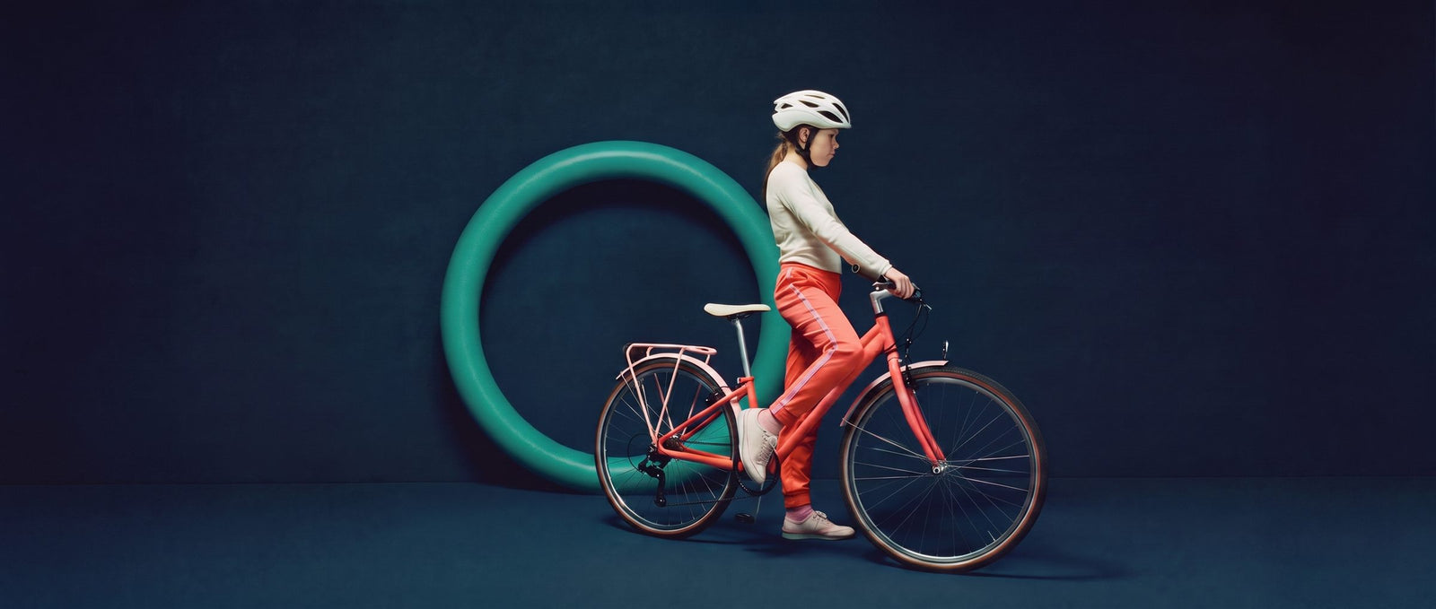 Tiener staat met een koraalrode 27,5 inch fiets voor een groene ring in een donkere navy studioset