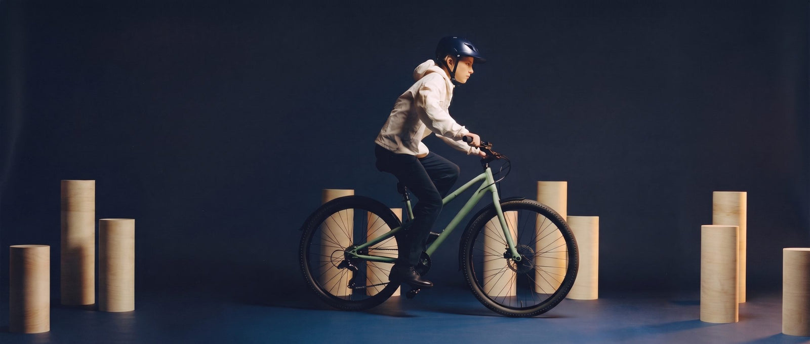 Tiener fietst op een zeegroene 29 inch fiets langs creme zuilen in een donkere navy studioset