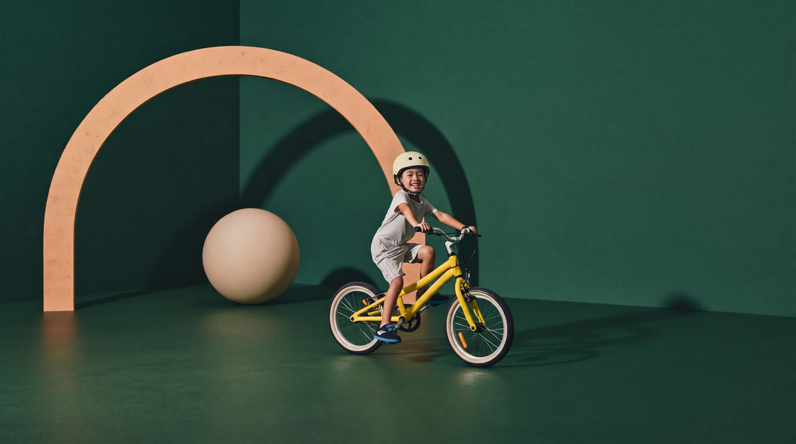 Trotse kleuter rijdt op een gele kinderfiets door een zeegroene studioset met perzikkleurige boog