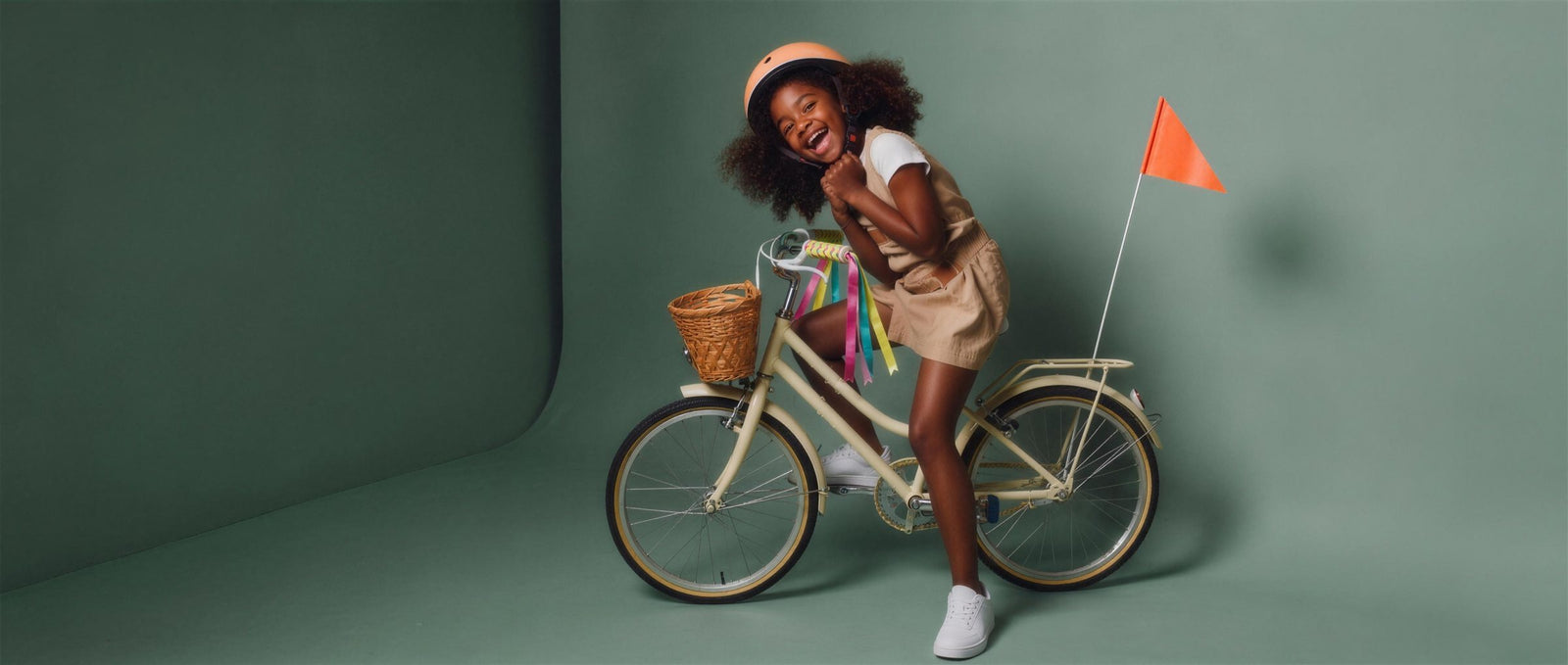 Lachend meisje klikt haar perzikkleurige helm vast op een cremekleurige fiets met rieten mandje, gekleurde linten aan het stuur en een oranje veiligheidsvlaggetje, in een groene studioset