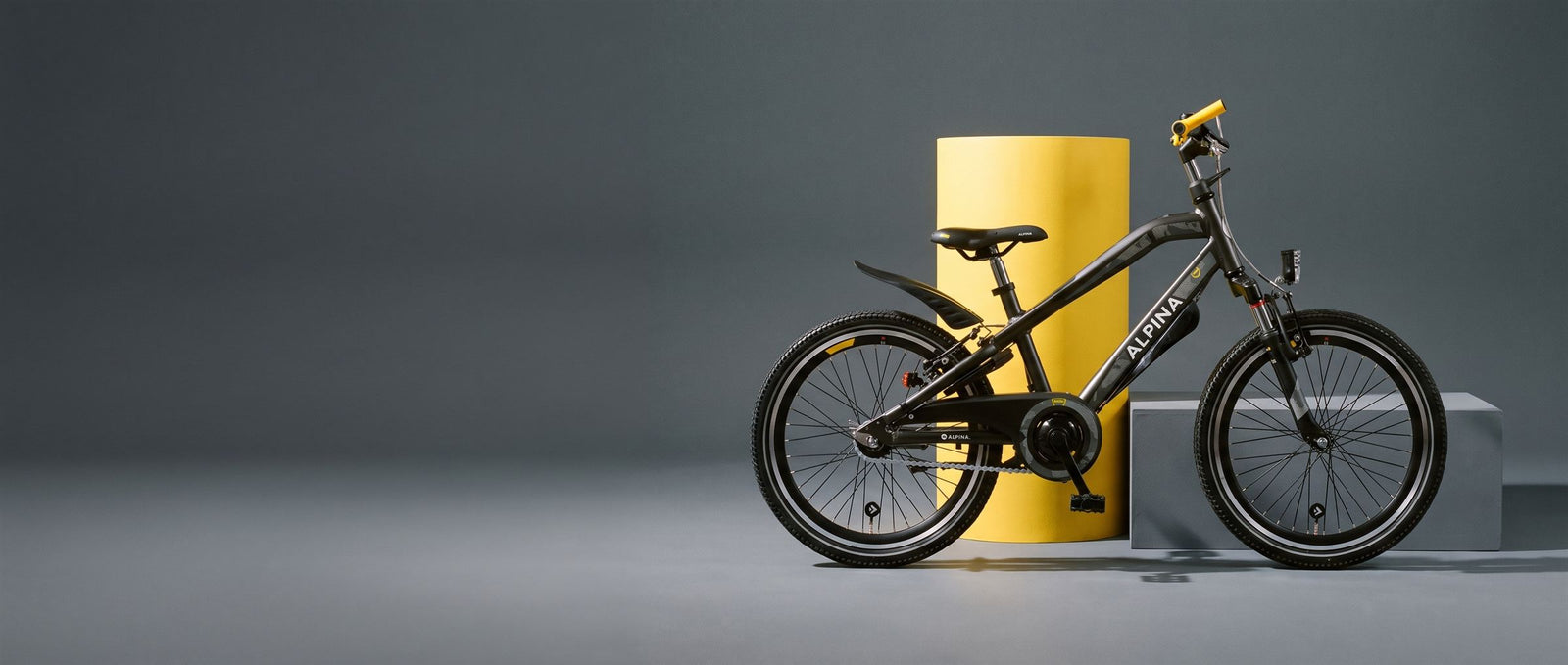 Antracietgrijze Alpina kinderfiets met gele grepen naast een gele zuil in een grijze studioset