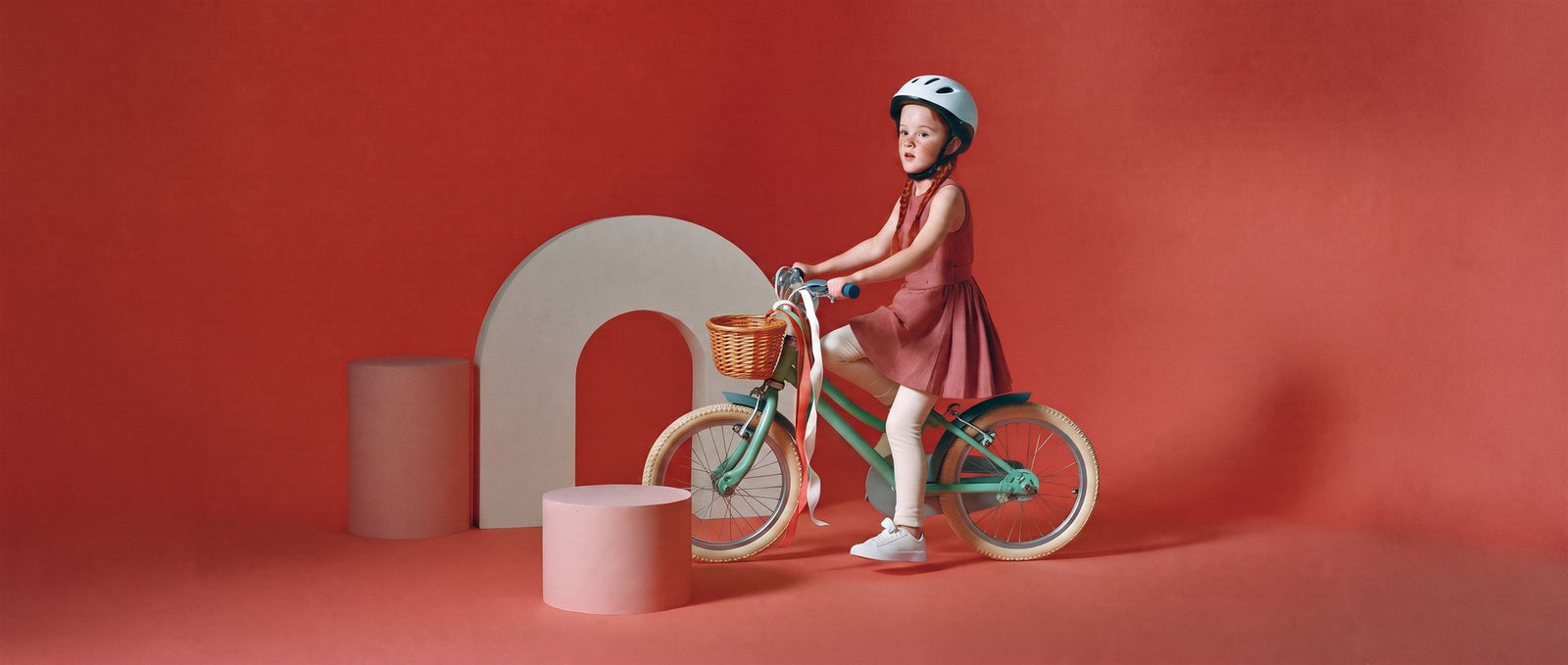Meisje met vlechtjes zit op een mintgroene fiets met mandje en linten in een koraalrode studioset
