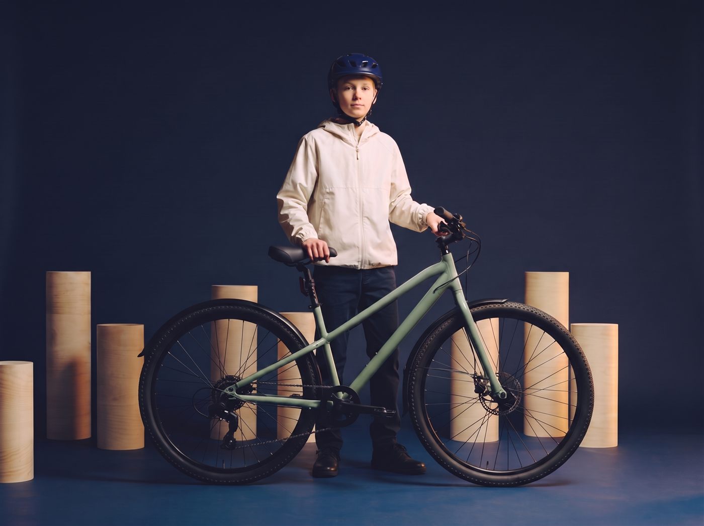 Tiener fietst op zijn zeegroene 29 inch fiets langs creme zuilen in een donkere navy studioset